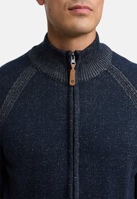 Marine gebreide trui met hoge kraag, voorzien van een volledige rits en leren trekband. Gestructureerd patroon met contrasterende lichte vlekken.