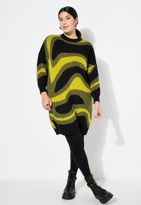 Tunic lavorata a maglia nera con audaci motivi a onda verde lime. Design oversize con polsini e orlo a coste. Abbinata a leggings neri e stivali chunky.