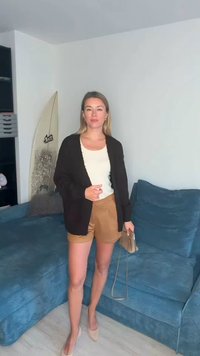 Musta neulejakku valkoisen paidan päällä, yhdistetty ruskeisiin shortseihin. Alastonväriset korkokengät ja pieni käsilaukku. Taustalla sininen sohva ja surffilauta.