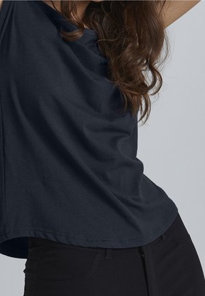 Kobieta z długimi kręconymi włosami ubrana w luźną ciemnogranatową koszulę i czarne spodnie na jednolitym szarym tle.