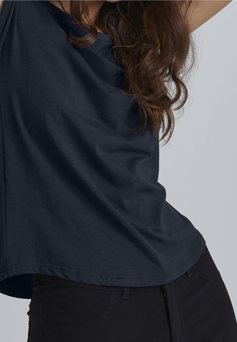 Femme aux longs cheveux bouclés portant une chemise ample bleu marine foncé et un pantalon noir sur un fond gris uni.