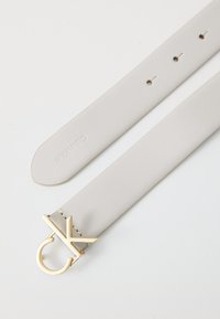 Curea Calvin Klein din piele gri deschis, cu o cataramă din metal aurit ce prezintă un design stilizat "CK". Textură netedă, cu striații pentru ajustare.