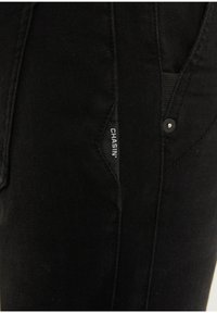 Close-up of black denim pants pocket with visible "CHASIN" label stitched inside the seam.