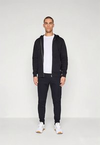 Μαύρο φούτερ με φερμουάρ πάνω από λευκό T-shirt, συνδυασμένο με μαύρη φόρμα και λευκά αθλητικά παπούτσια με καφέ σόλες. Απλός σχεδιασμός, χωρίς σχέδια.