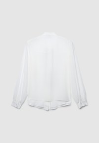 Blusa bianca a maniche lunghe con polsini con bottoni, dettaglio della piega sul retro e orlo leggermente curvato su uno sfondo grigio chiaro.