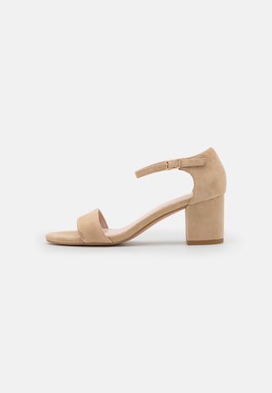 Beige ruskind åben tå sandal med blokhæl og ankelrem med spænde på en ensfarvet lys baggrund.