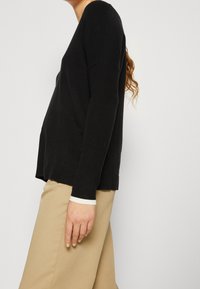 Maglione nero con maniche lunghe, tessuto leggero e vestibilità rilassata. Presenta una scollatura a V e spacchi ai lati dell'orlo.