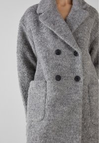Grå ullblandad dubbelknäppt kappa med stora svarta knappar, snibbkrage och sidofickor; den strukturerade tyget ger värme.
