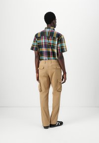 Short-sleeve plaid shirt in black, yellow, and teal; tan cargo pants with large side pockets; black sneakers. Viewed from the back.