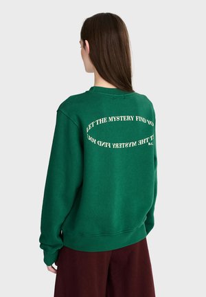 Personne aux longs cheveux bruns portant un sweat-shirt vert avec un texte crème incurvé dans le dos, associé à un pantalon rouge foncé, vue de dos.