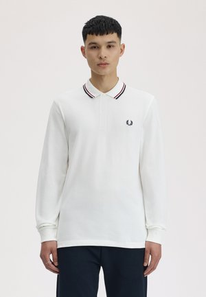 Ung mand iført en hvid langærmet polo med stribet krave og sorte bukser, stående mod en ensartet lys baggrund.