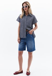 Svart och vit rutad kortärmad skjorta, denim Bermuda-short med fransar, röda platta skor och vita solglasögon. Minimalistisk stil.