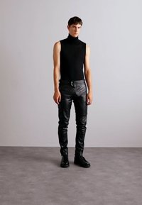 Črna brezrokavna majica s turtleneck izrezom, v kombinaciji s tesnimi črnimi usnjenimi hlačami in črnimi čevlji, stoji na nevtralnem sivo tlorisu.