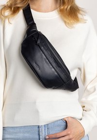 Černá kožená ledvinka s zipovým kapsou, elegantní design, zakřivený tvar a nastavitelné rameno nošené přes bílý svetr.