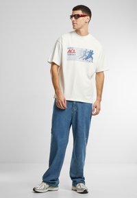 White cotton t-shirt featuring a blue and red graphic print. Worn with loose blue jeans and silver sneakers, accessorized with red sunglasses.
