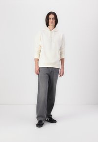 Hanorac crem cu buzunar tip kangaroo, asortat cu blugi gri, largi. Outfitul este completat cu adidași negri cu accente albe.