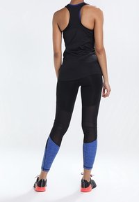 Černý atletický top s racerback střihem. Černé legíny s texturovanými modrými spodními panely a černými doplňky. Tmavé tenisky s červenými podrážkami.