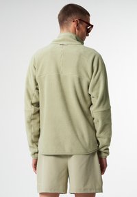 Hellgrüner Fleece-Pullover mit hohem Kragen, strukturiertem Oberflächenmaterial und Rückennahtdetails, kombiniert mit passenden kurzen Hosen.