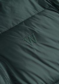 Tissu matelassé vert foncé avec un logo "W" brodé et une fermeture éclair partiellement visible sur le côté gauche.