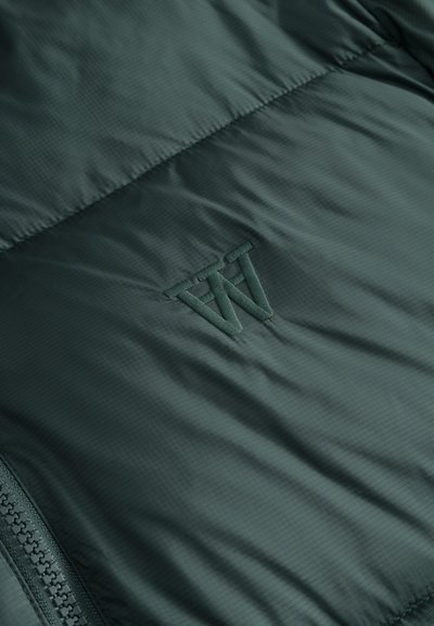 Tissu matelassé vert foncé avec un logo "W" brodé et une fermeture éclair partiellement visible sur le côté gauche.
