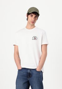 White cotton t-shirt with a round neck and short sleeves, featuring a black logo print on the chest. Paired with blue denim jeans and a green cap.