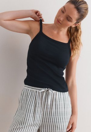 Kvinde justerer sort top med spaghetti-stropper, iført hvide og grå lodrette stribede bukser med snøre, foran en ensartet lys baggrund.