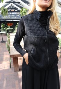 Melnā, bezrocīgā veste ar faktūrētu augšējo daļu un mīkstu, vilnas līdzīgu apakšdaļu; rāvejslēdzējs un divas priekšējās kabatas.