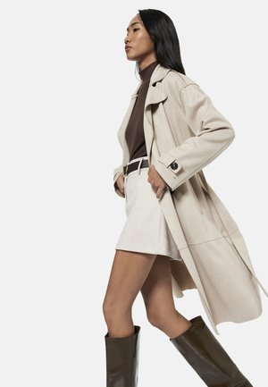 Kvinde med langt sort hår iført beige trenchcoat, brun rullekrave, beige nederdel, bælte og høje mørkebrune støvler, går foran en ensfarvet baggrund.