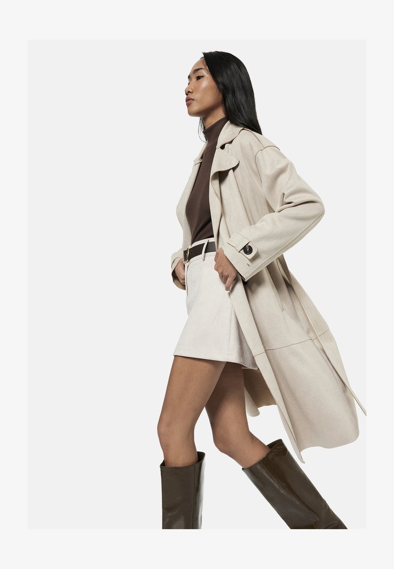 Frau mit langen schwarzen Haaren trägt beige Trenchcoat, braunen Rollkragenpullover, beigen Rock, Gürtel und hohe dunkelbraune Stiefel und geht vor einfarbigem Hintergrund.