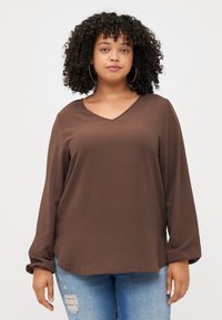 Une jeune femme aux cheveux bouclés porte un haut brun à col en V et un jean bleu déchiré, souriant doucement devant un fond gris uni.
