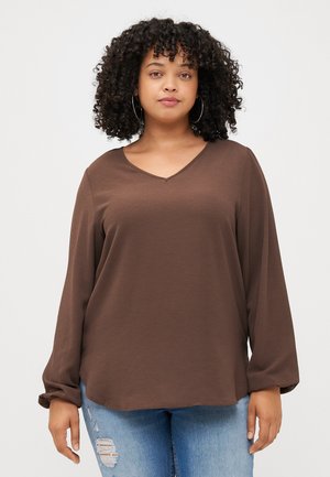 Une jeune femme aux cheveux bouclés porte un haut brun à col en V et un jean bleu déchiré, souriant doucement devant un fond gris uni.