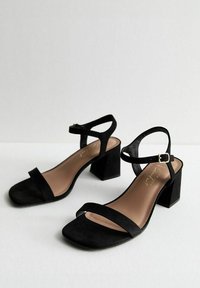 Sandalias de tacón bloque de ante negro con una única tira en el dedo y una correa en el tobillo con un cierre dorado. Plantilla suave con una suela beige.