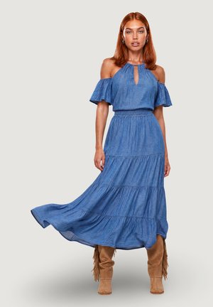 Donna con capelli rossi che indossa un vestito blu con scollo a halter e stivali di pelle scamosciata color cammello con frange, in piedi davanti a uno sfondo chiaro e neutro.