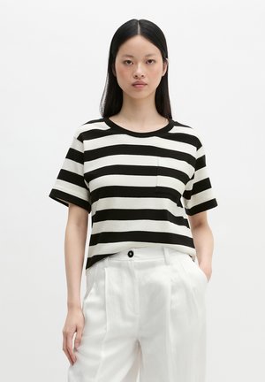 Femme aux longs cheveux noirs portant un t-shirt à manches courtes rayé horizontal noir et blanc et un pantalon blanc taille haute, debout avec une main dans la poche.