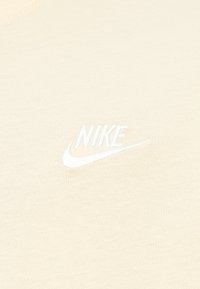 Ανοιχτό μπεζ T-shirt από μαλακό ύφασμα, με λευκό κεντημένο λογότυπο Nike στην αριστερή πλευρά. Ομαλή υφή με απλό σχέδιο.