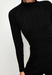 Woman wearing a fitted black ribbed turtleneck sweater and high-waisted black pants against a plain light background.