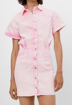 Kobieta w jasno-różowej, dżinsowej sukience z krótkim rękawem, zapinanej na guziki, z kołnierzykiem i przednimi kieszeniami, stojąca na prostym tle.