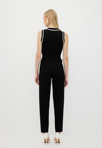 Jumpsuit sans manches noir avec des accents blancs, taille cintrée et jambes effilées. Porté avec des chaussures à talons hauts de couleur crème.