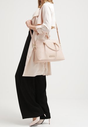 Lyserød tekstureret håndtaske med to håndtag og en aftagelig rem, præsenteret ved siden af en person iført en lang, beige trenchcoat og sorte bukser.
