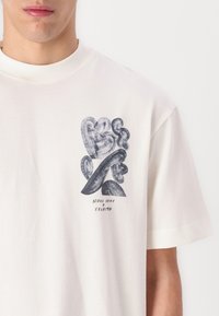 Medvilninis marškinėliai off-white spalvos, su tamsiai mėlyna grafika. Meninė kompozicija apima abstrakčius formas su tekstu žemiau šiuolaikišku šriftu.