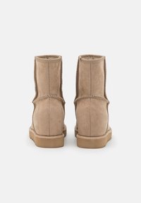 Paire de bottes d'hiver en daim beige avec couture visible et semelles en caoutchouc plates, vues de dos sur fond blanc.