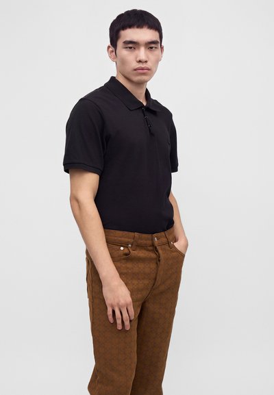 Jeune homme aux cheveux courts noirs portant un polo noir et un pantalon marron à motifs, debout avec une main dans la poche sur un fond uni.