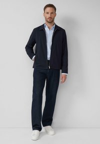 Marinejacke mit Reißverschluss und spitzem Kragen, kombiniert mit einem hellblauen Hemd und dunklen Denimhosen. Weiße Sneaker runden das Outfit ab.
