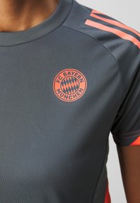 Camiseta atlética gris con el logo del FC Bayern München en naranja. Los detalles notables incluyen rayas naranjas en los hombros y un material texturizado.