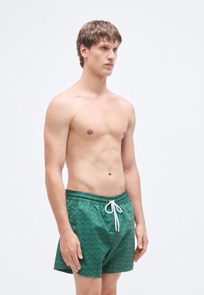 Πράσινα μαγιό σορτς με ζιγκ-ζαγκ μοτίβο, κατασκευασμένα από ελαφρύ υλικό, με ελαστική μέση και λευκό κορδόνι.
