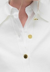 Bílá košile s texturovaným povrchem, vybavená třemi zlatými knoflíky a malou zlatou broží na límci.