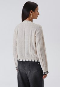 Maglione lavorato a maglia color crema con motivi a treccia e orlo nero a coste, dal design cropped, con maniche larghe e una finitura strutturata.