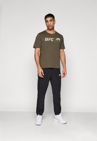 Oliven grøn t-shirt med korte ærmer og UFC-logo, parret med sorte træningsbukser. Modellen har hvide sneakers på. Glat stof, afslappet pasform.
