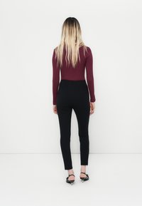 Donna con lunghi capelli biondi che indossa un top lungo bordeaux, pantaloni neri aderenti e scarpe con tacco nere, di fronte a uno sfondo bianco.