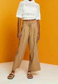 Beige wide-leg trousers with front pleats and drawstring waist. Worn with a cropped white open-knit top. Background is a solid orange.